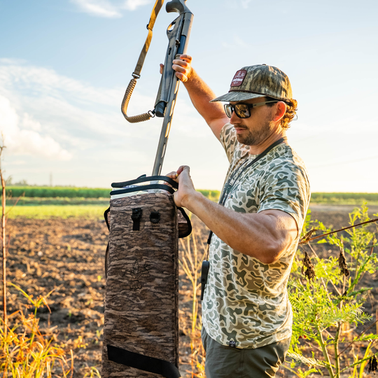 Man in camouflage gear holding a rifle and bag in an open field