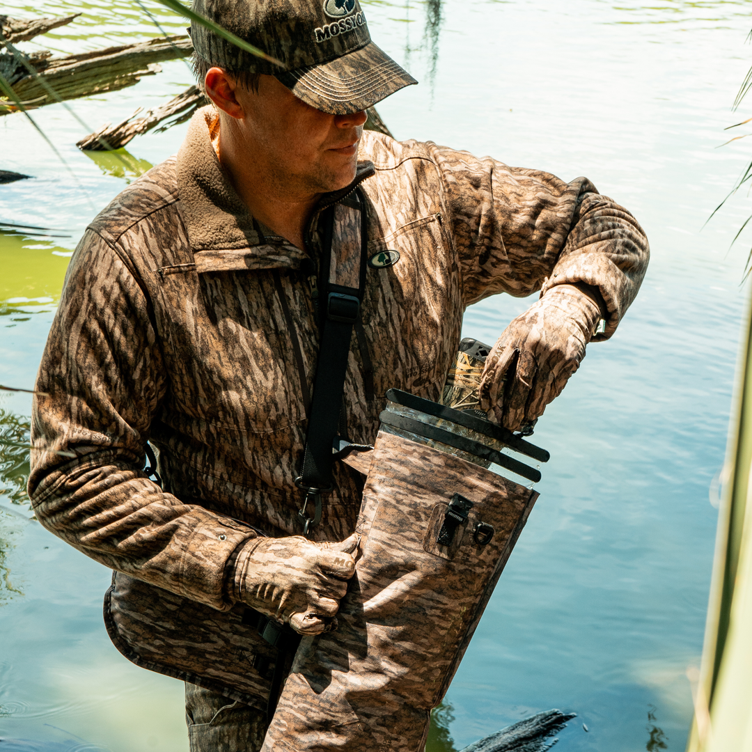 Man in camouflage clothing and waders standing in water, holding a fishing rod.