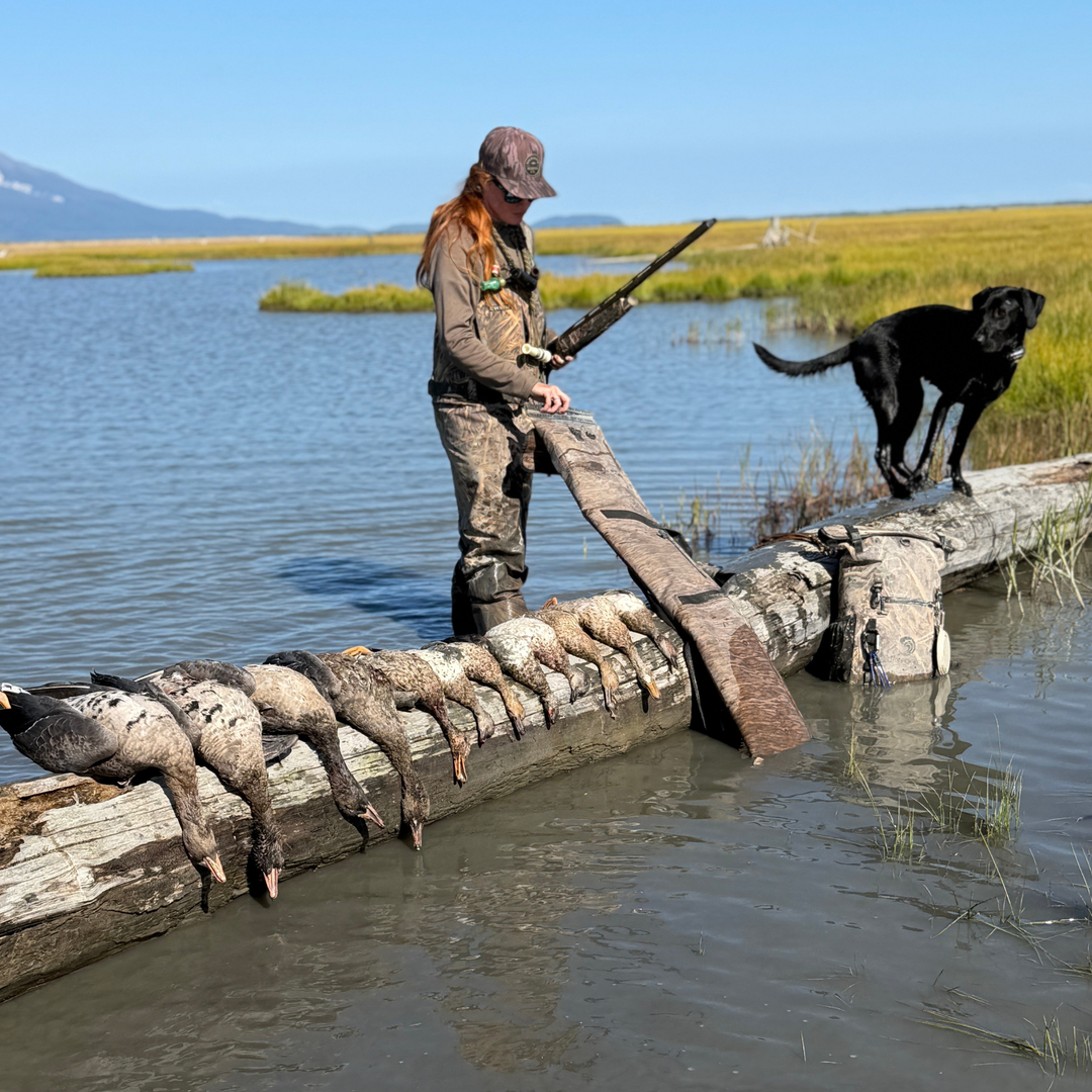 A woman in camouflage stands in shallow water holding a shotgun while balancing a long plank log loaded with hunted ducks. A black dog stands nearby on the log in an outdoor marsh setting under a blue sky.